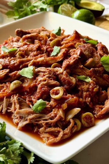 Slow cooker Spanish pulled pork on a serving plate with green olives and cilantro.