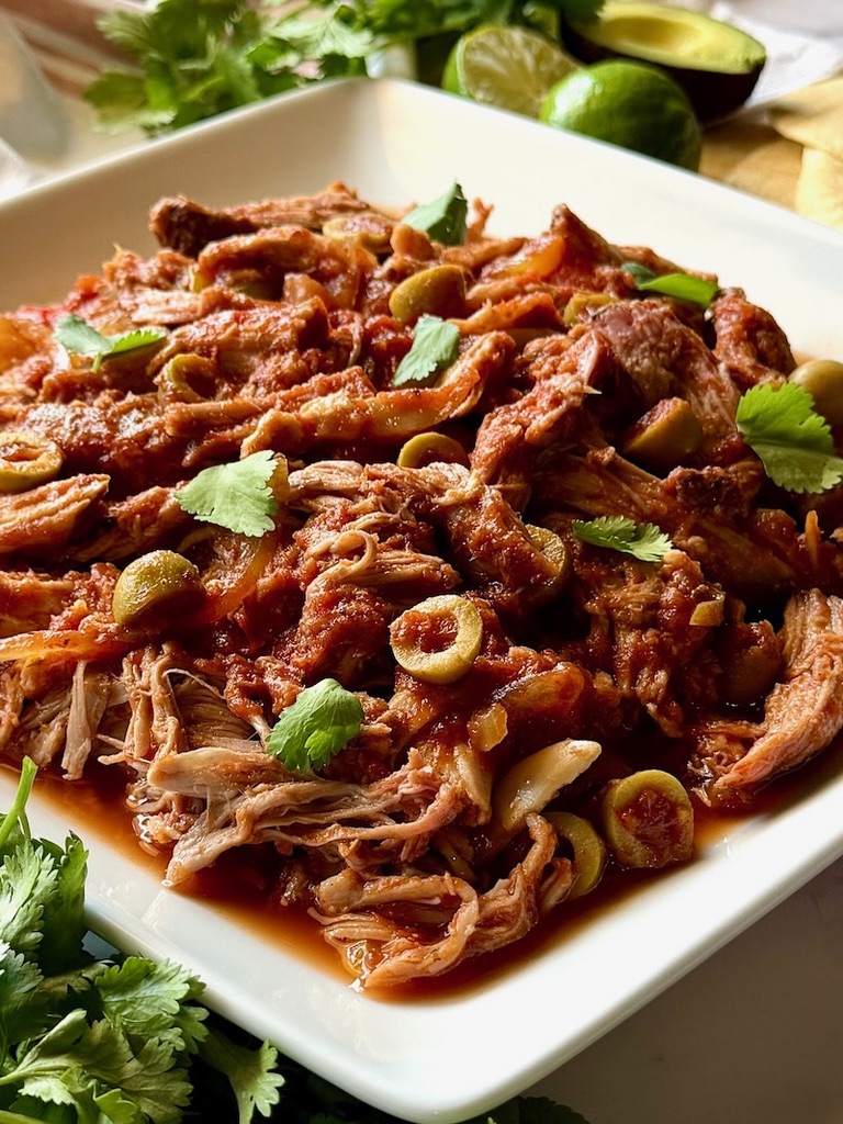 Slow cooker Spanish pulled pork on a serving plate with green olives and cilantro.