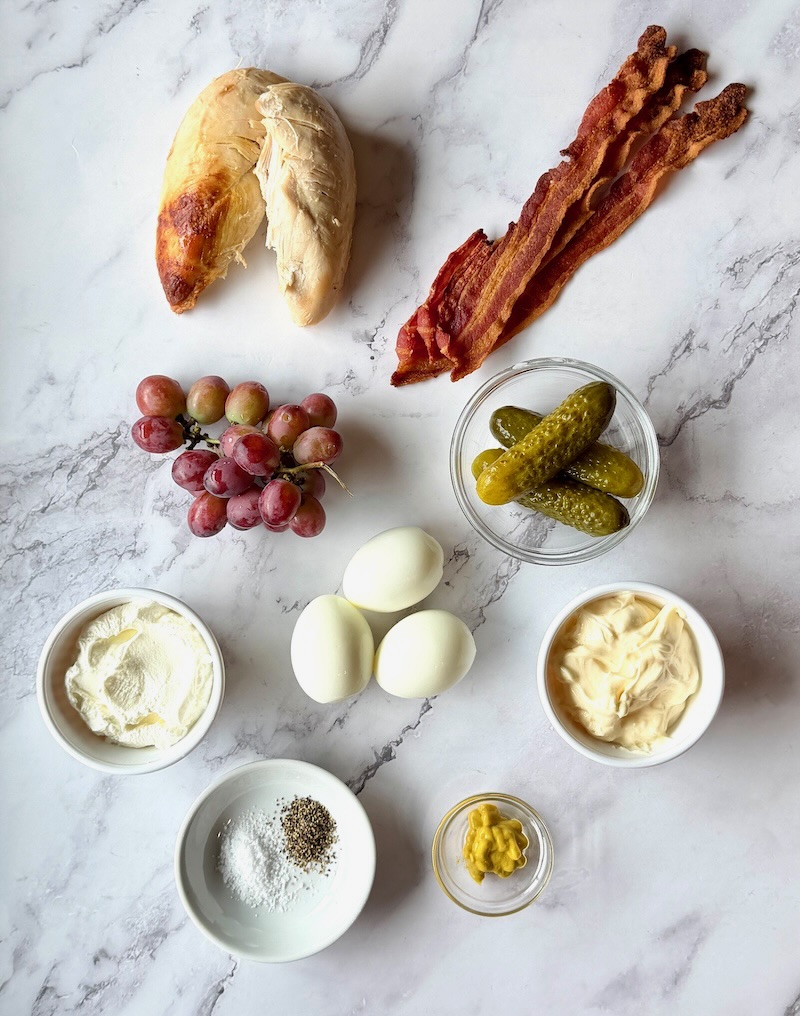 Ingredients displayed for Easy chicken salad with bacon. Rotisserie chicken, crispy bacon, red grapes, dill pickles, hard boiled eggs, Greek yogurt, mayo, yellow mustard, salt and pepper.