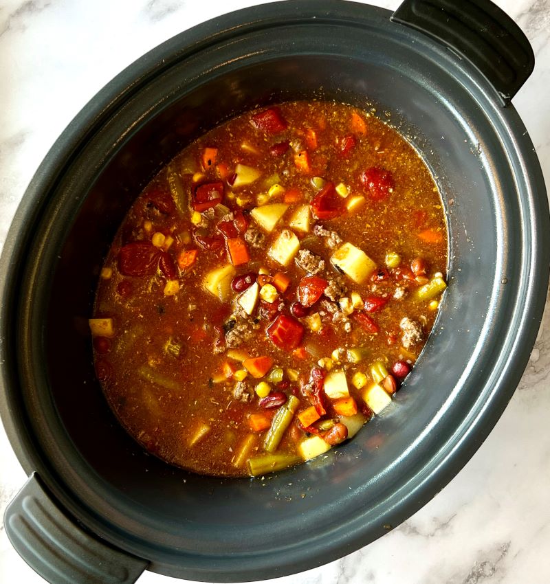 To the ground beef, onions and garlic in slow cooker, add beans, potatoes, carrots, corn, seasonings, fire roasted tomatoes, and beef stock.