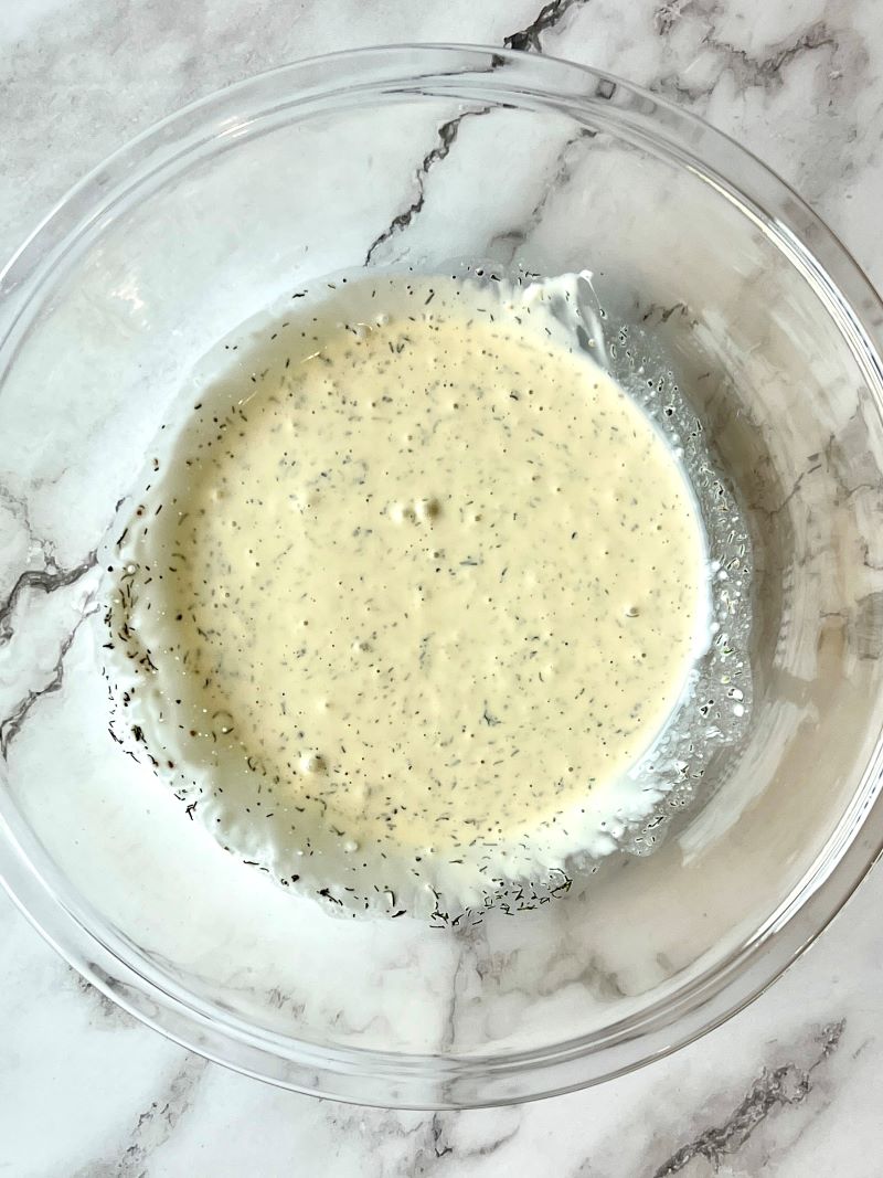 Dressing ingredients combined in a bowl. Mayo, Greek yogurt, sugar, dried dill, salt, pepper, apple cider vinegar.