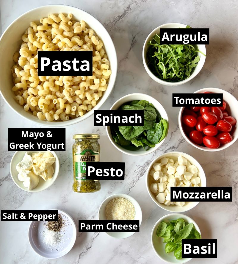 Ingredients for creamy pesto pasta salad. Spiral pasta, tomatoes, spinach, arugula, mozzarella, mayo, Greek yogurt, pesto, parmesan cheese, salt, pepper, and basil.