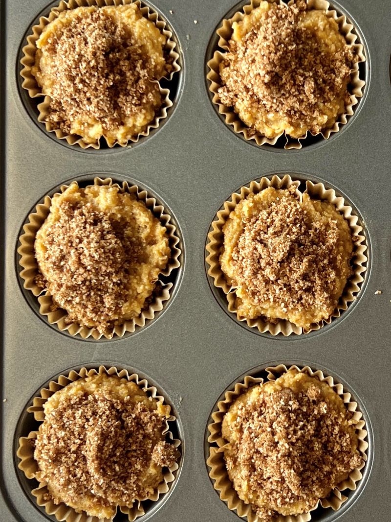 Cinnamon roll breakfast muffins layered in a muffin pan with muffin liners. Muffin batter, cinnamon crumble, more muffin batter, then top with more crumble. 