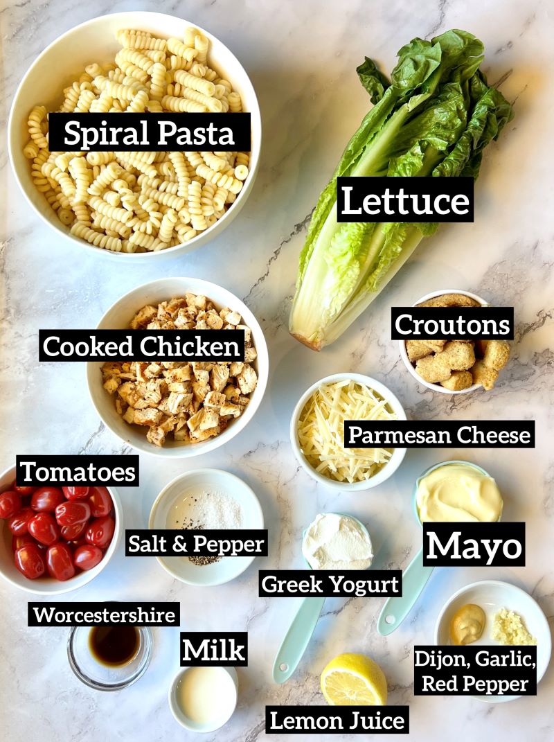 Ingredients arranged out on the counter for chicken Caesar pasta salad, include cooked spiral pasta, cooked chicken breast, romaine lettuce, croutons, parmesan cheese, cherry tomatoes, mayo, Greek yogurt, Worcestershire sauce, milk, lemon juice, Dijon mustard, garlic, red pepper flakes, salt and pepper. 