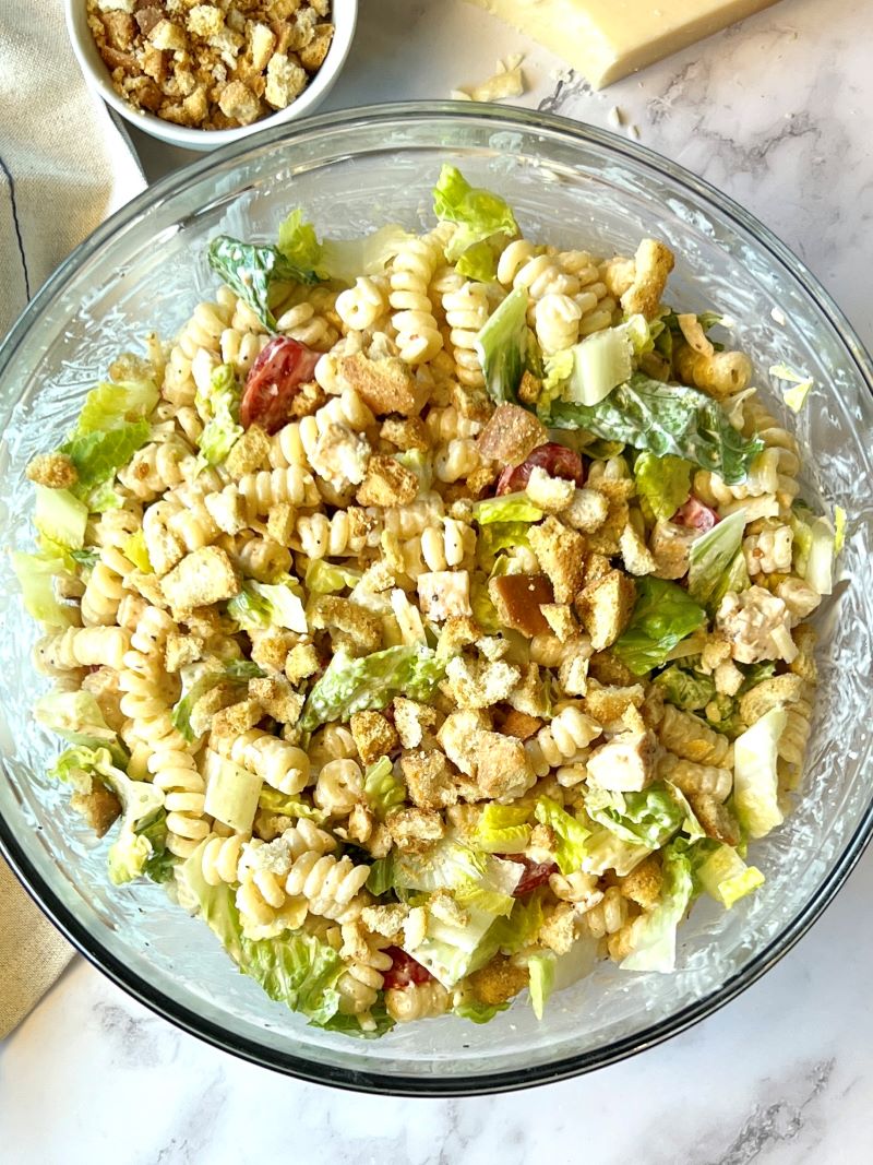 Cooked pasta in a large bowl tossed with cooked chicken, tomatoes, lettuce, parmesan cheese, Caesar dressing, and topped with croutons. 