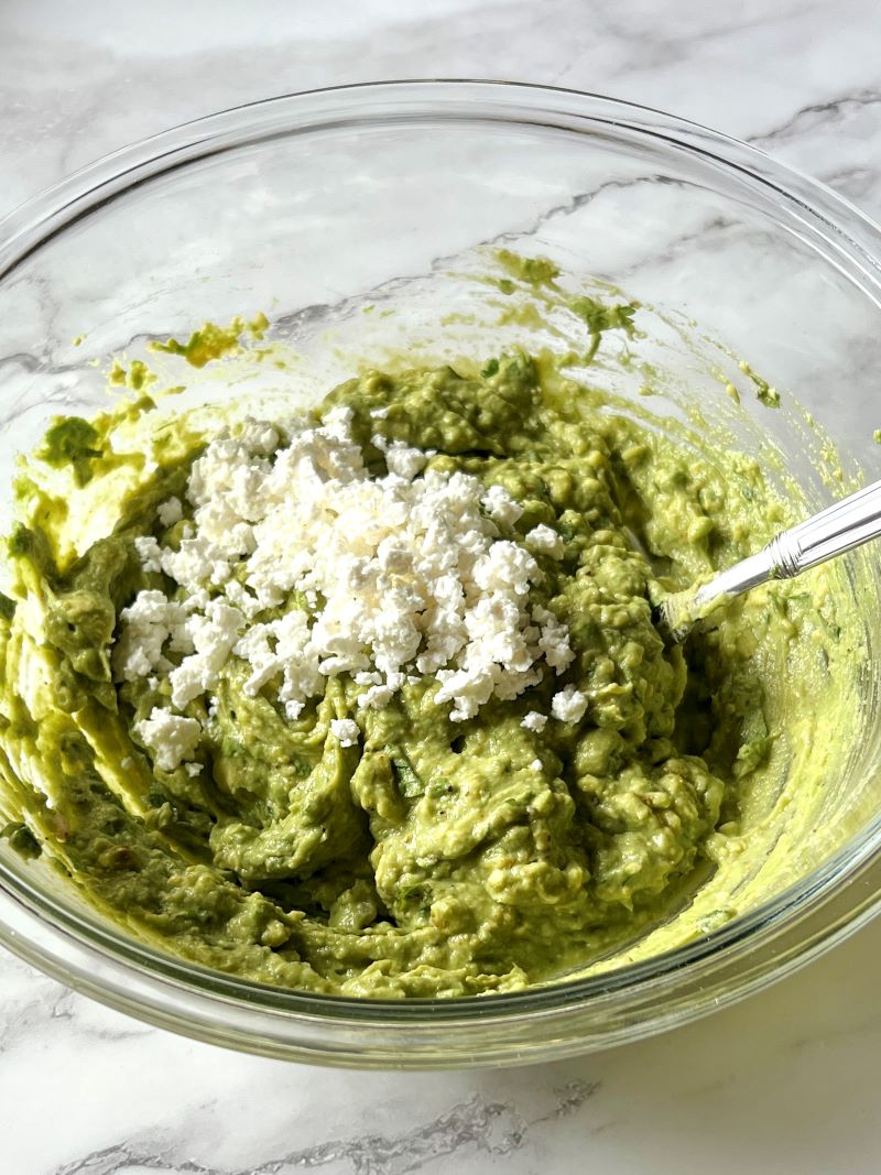 Mashed avocados with lime zest, juice, onion, cilantro, ground cumin, cayenne pepper, salt, pepper, and feta cheese in a bowl with a fork.