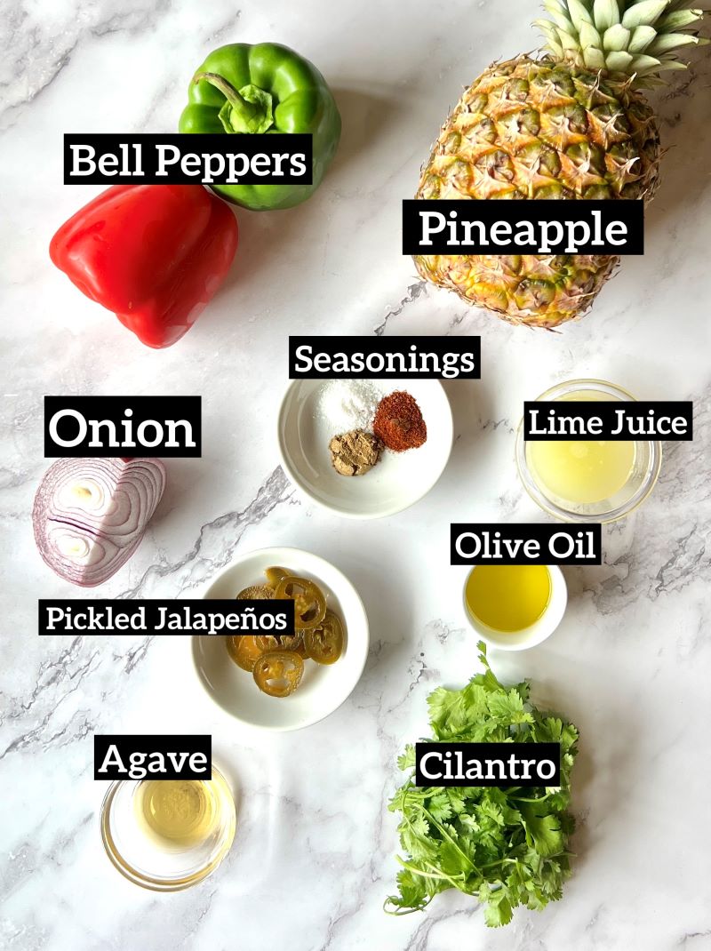 Ingredients displayed for fresh pineapple salsa. Green and red bell peppers, red onion, fresh pineapple, lime juice, spices, olive oil, pickled jalapenos, agave, and cilantro. 