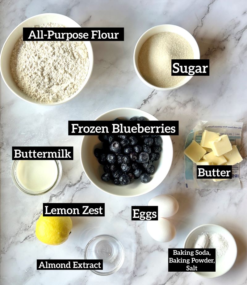 Ingredients for blueberry buckle set out. Flour, baking soda, baking powder, lemon zest, sugar, butter, eggs, almond extract, buttermilk, frozen blueberries. 
