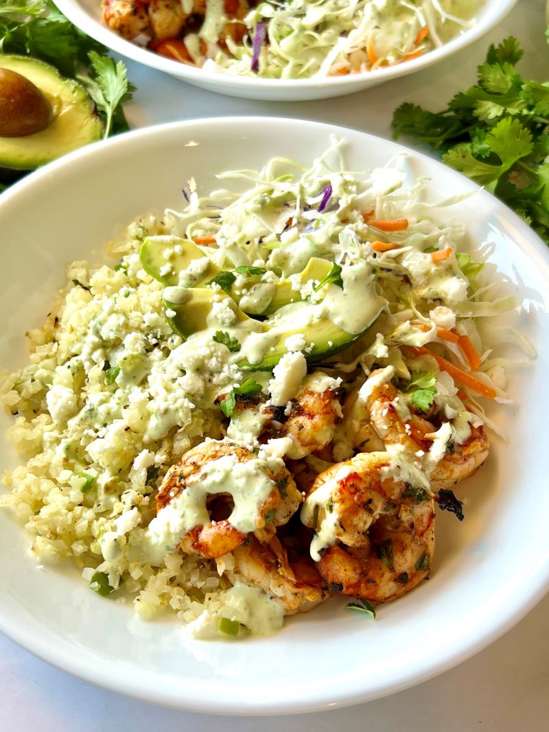 Grilled shrimp bowls assembled in a bowl with cauliflower rice, shrimp, cabbage, avocado, and dressing.