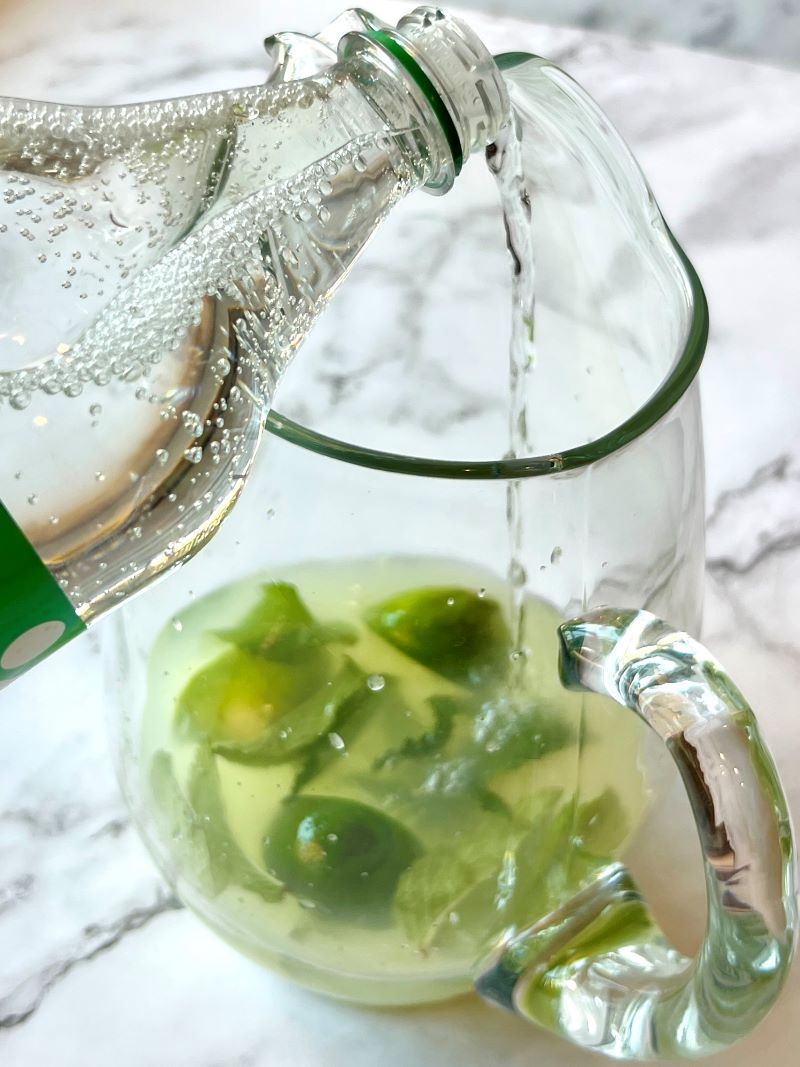 Adding sprite to a glass pitcher with lemonade mix, limes and mint.