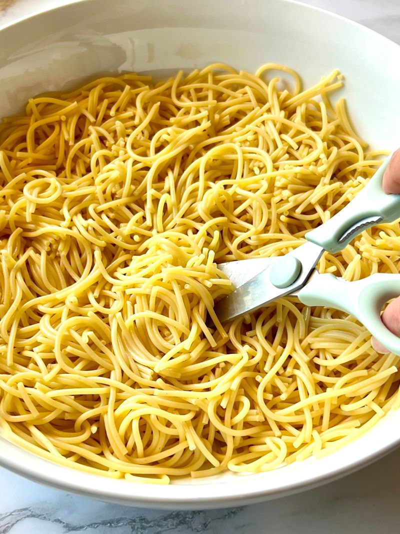 Large bowl with cooked spaghetti, using kitchen shears to cut the noodles smaller.