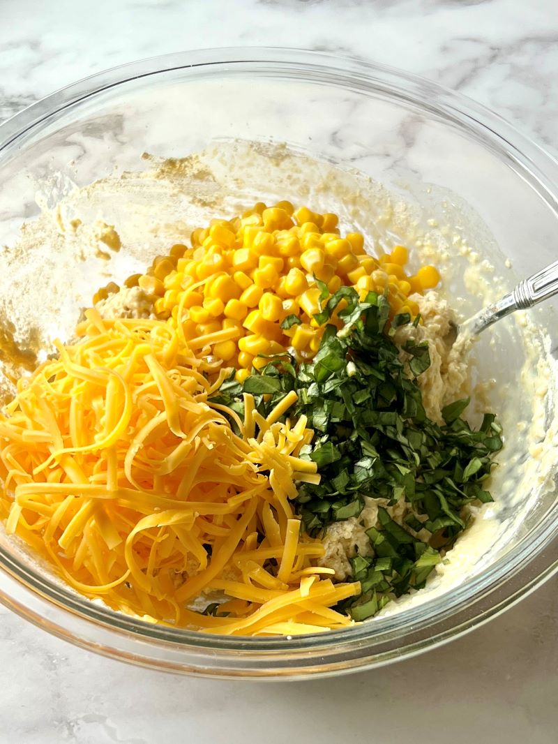 Muffin batter with shredded cheese, corn, and basil in a bowl.