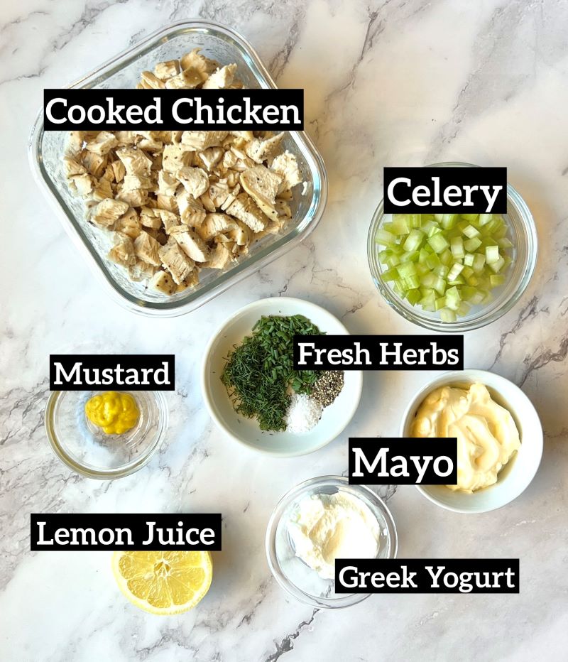 Ingredients displayed for simple chicken salad. Cooked chicken breast cut in chunks, celery, mayo, mustard, Greek yogurt, fresh herbs, salt, pepper, and lemon juice. 