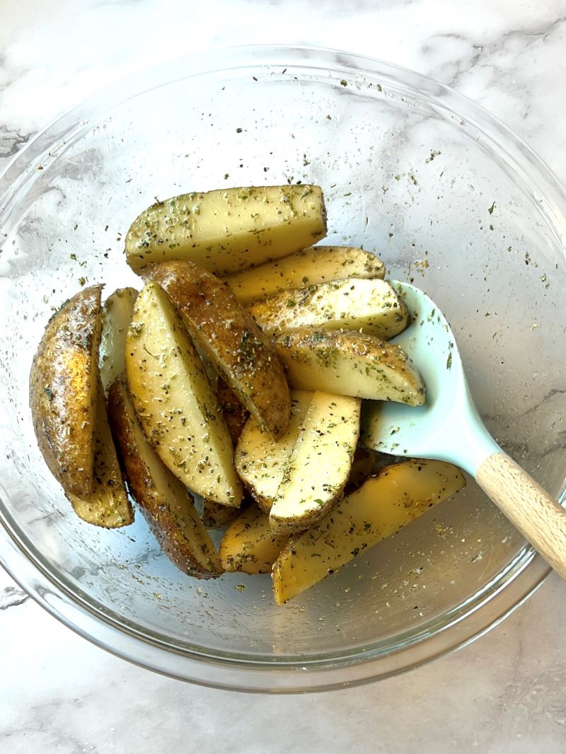 Potato wedges combined with olive oil and seasonings in a bowl with a spoon
