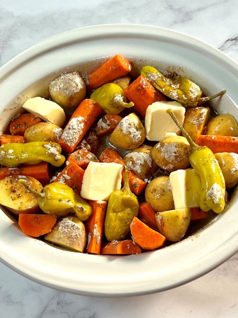 Roast added to the slow cooker and topped with carrots, potatoes, Au jus, ranch mix, pepperoncini peppers, beef broth, and butter.