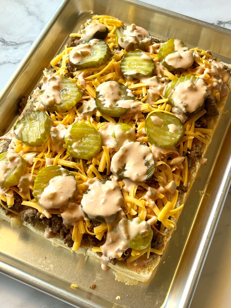 Bottom bun layered with sauce, ground beef, cheddar cheese, pickle chips, and more sauce.