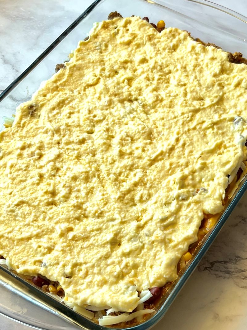 Casserole layered in a baking dish with meat filling, cheese, and cornbread mix.