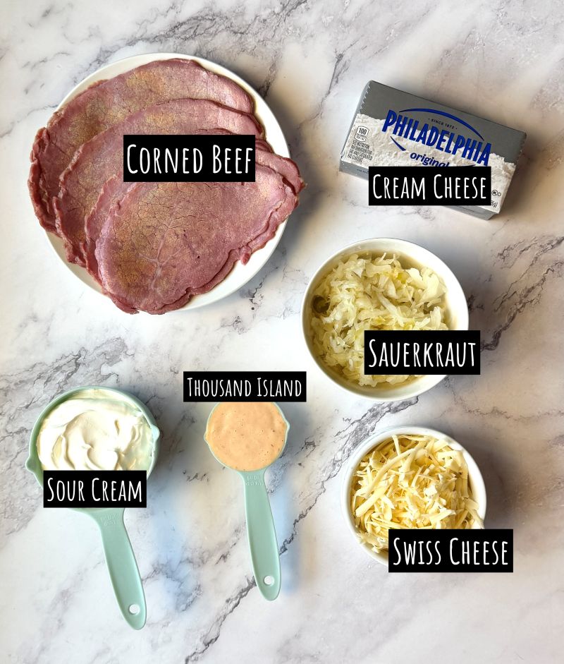 Ingredients displayed for slow cooker Reuben dip. Corned beef, cream cheese, sauerkraut, thousand island dressing, sour cream, Swiss cheese. 