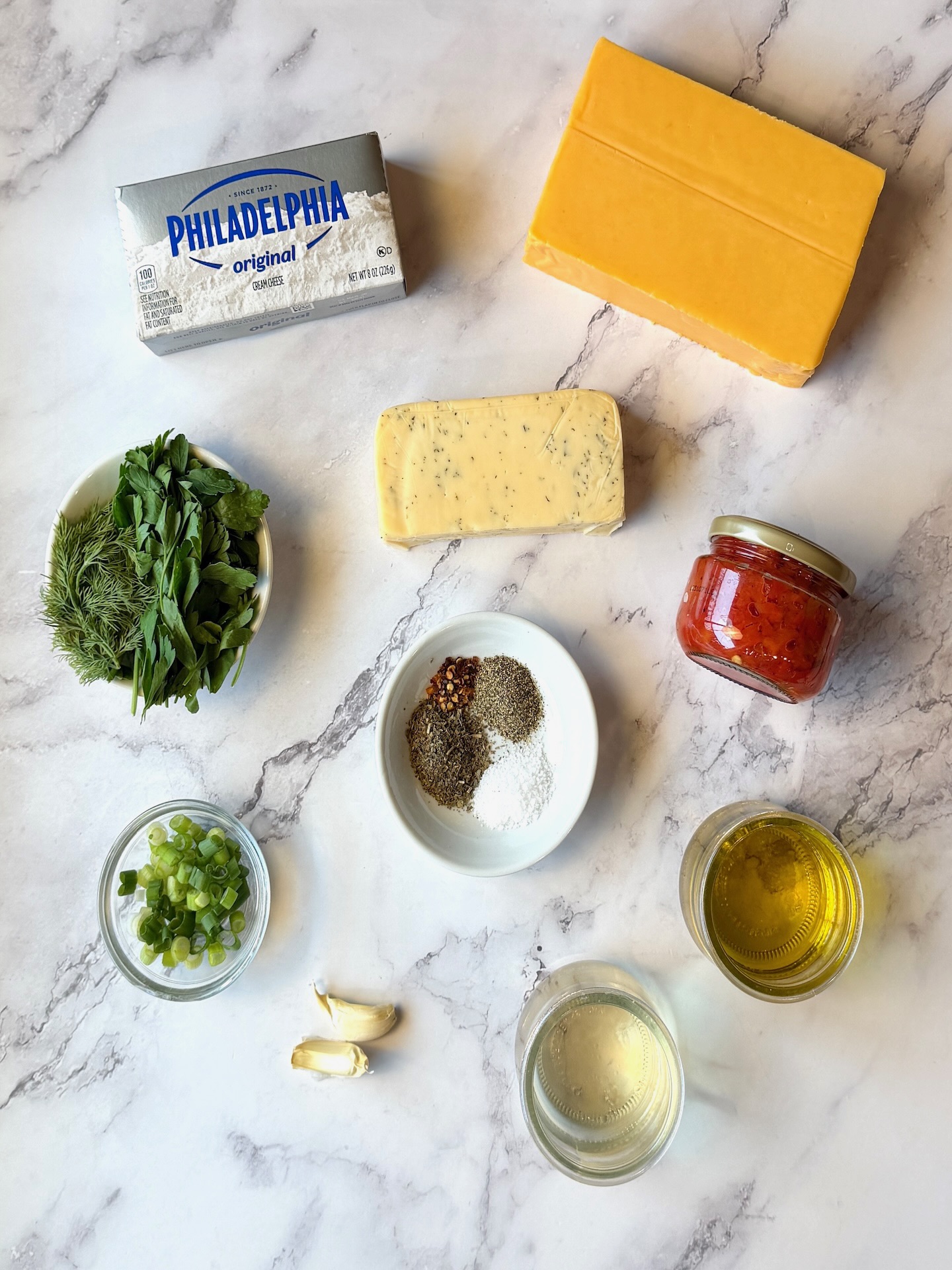 Ingredients displayed for marinated cheese. Cheddar cheese block, havarti cheese block, cream cheese block, jarred pimentos, fresh herbs, seasonings, garlic, olive oil, vinegar, and green onions.