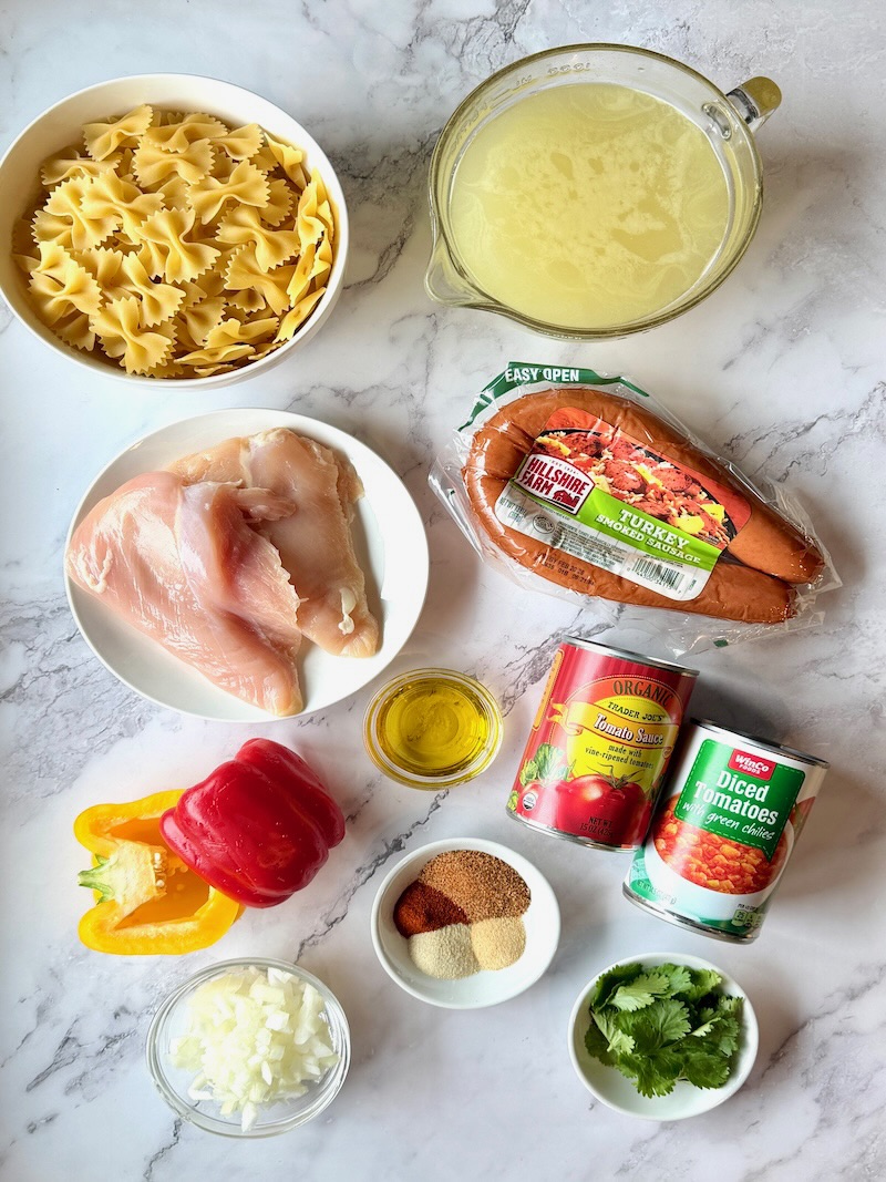 Ingredients displayed for one-pot jambalaya pasta. Bow tie pasta, chicken broth, chicken breast, smoked sausage, olive oil, tomato sauce, tomatoes and green chils, seasonings, onions, bell peppers, and cilantro. 