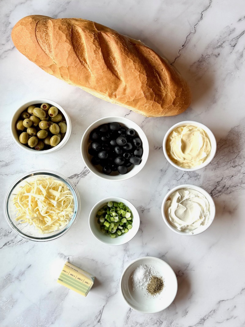 Ingredients displayed for cheesy olive bread. French bread, black and green olives, mayo, sour cream, Jack cheese, green onions, butter, salt and pepper.