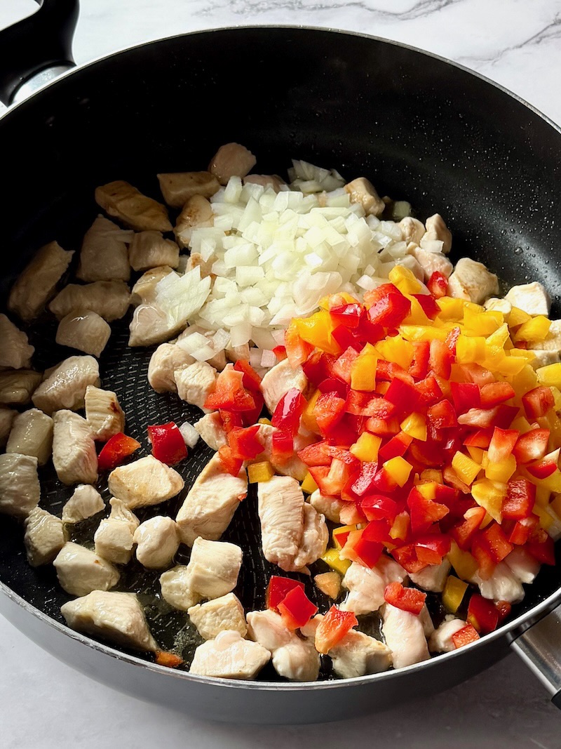 Cooked chicken pieces with peppers and onions in a large skillet. 
