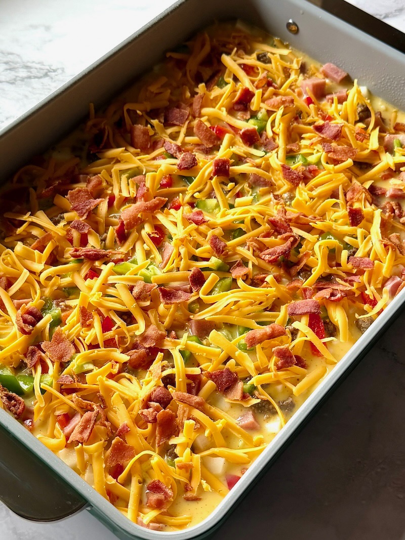 Layered casserole with hash browns, sausage, ham, peppers, egg mixture, cheese, and cooked bacon. 