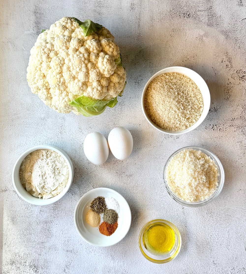 Ingredients displayed for crunchy parmesan roasted cauliflower. Raw cauliflower head, eggs, panko bread crumbs, flour, parmesan cheese, seasonings, and olive oil. 
