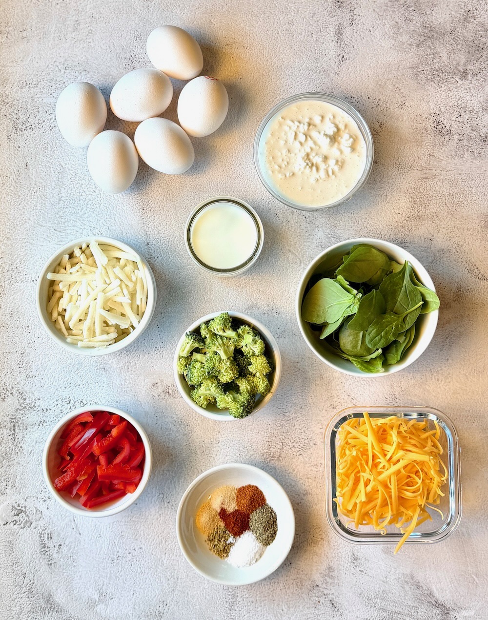 Ingredients displayed for veggie breakfast casserole. Eggs, cottage cheese, milk, fresh spinach, fresh broccoli, red bell peppers, frozen hash browns, seasonings, and cheddar cheese. 