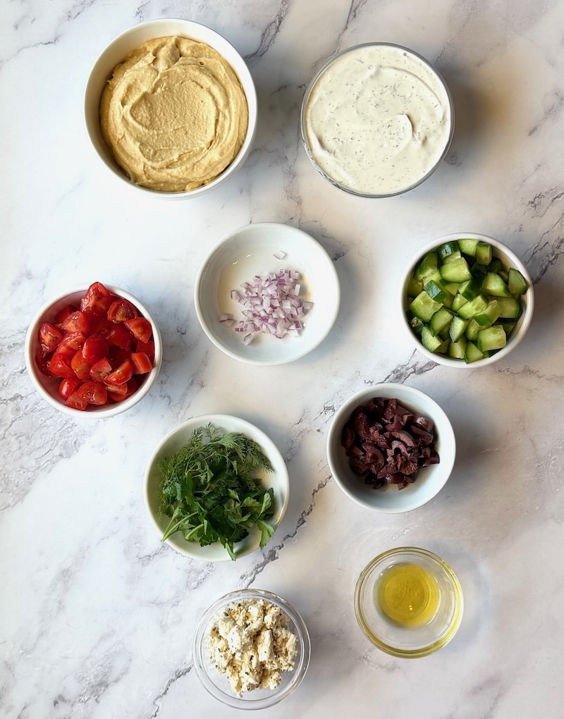 Ingredients displayed for easy Greek layered dip. Hummus, tzatziki, red onion, tomatoes, cucumbers, olives, feta cheese, fresh dill, parsley, and olive oil.
