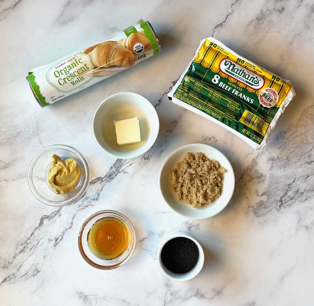 Ingredients displayed for sweet glazed pigs in a blanket. Organic Crescent rolls, Nathan's beef hot dogs, butter, brown sugar, Dijon mustard, honey, poppy seeds.
