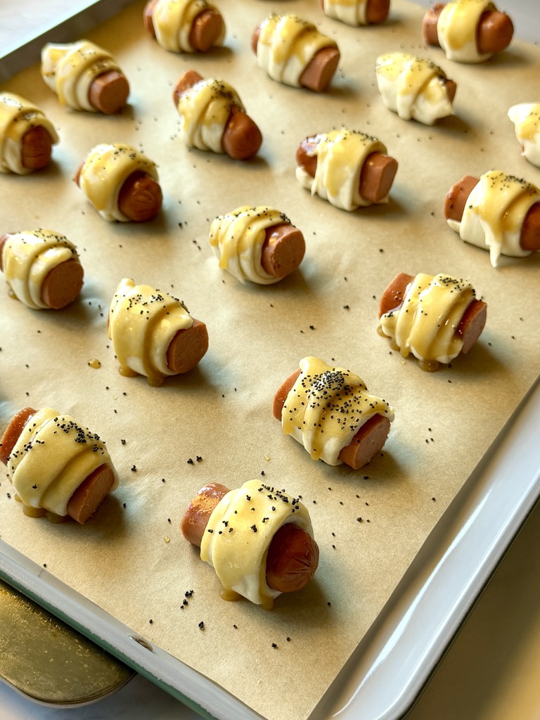 Pigs in a blanket unbaked on a sheet pan with glaze and topped with poppy seeds.