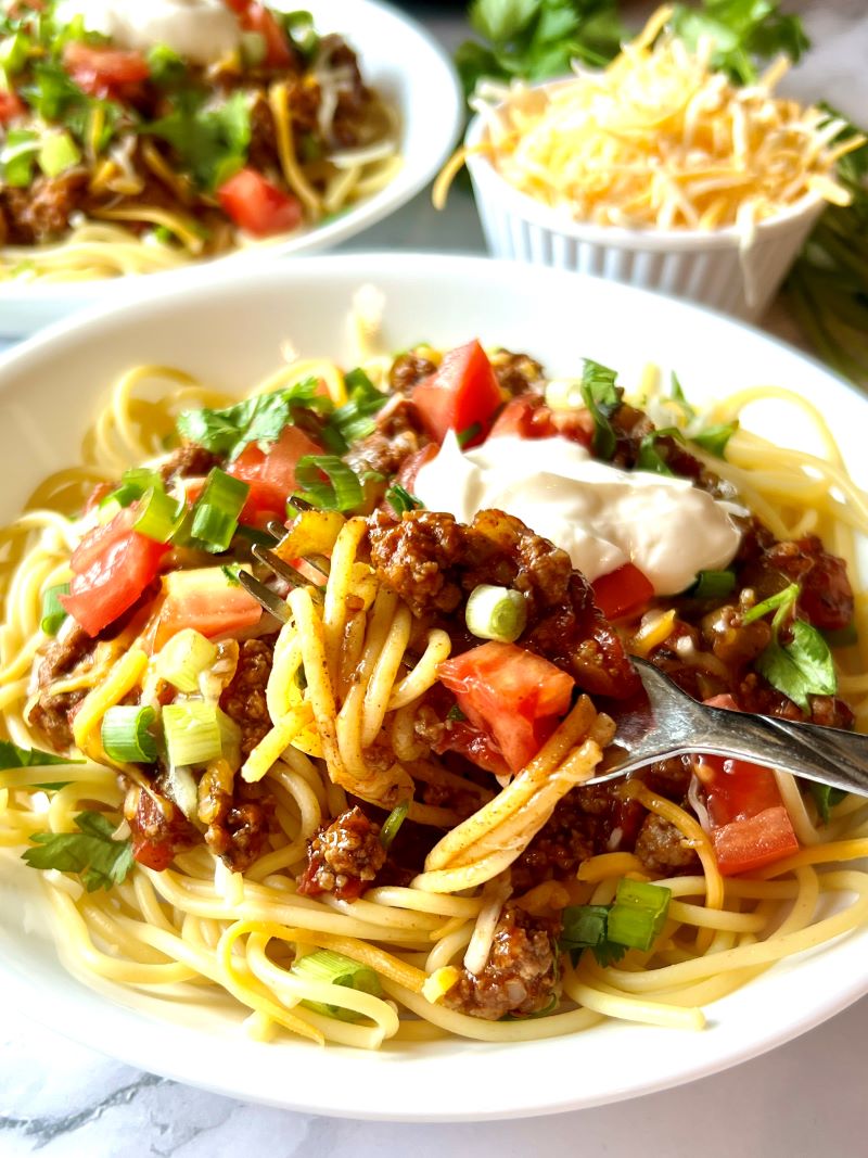 Bowl of taco spaghetti topped with cheese, sour cream, tomatoes, and green onions.