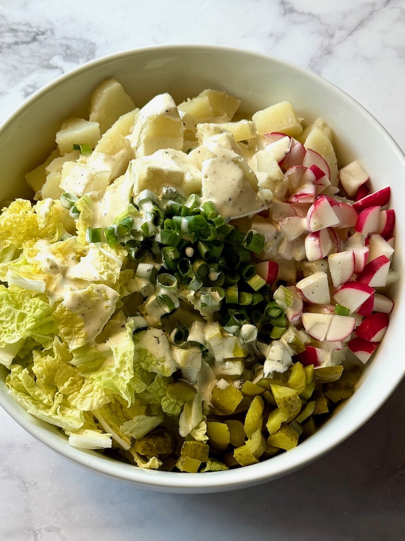 Potatoes in a bowl with cabbage, radishes, pickles, green onions, and dressing for Irish potato salad.