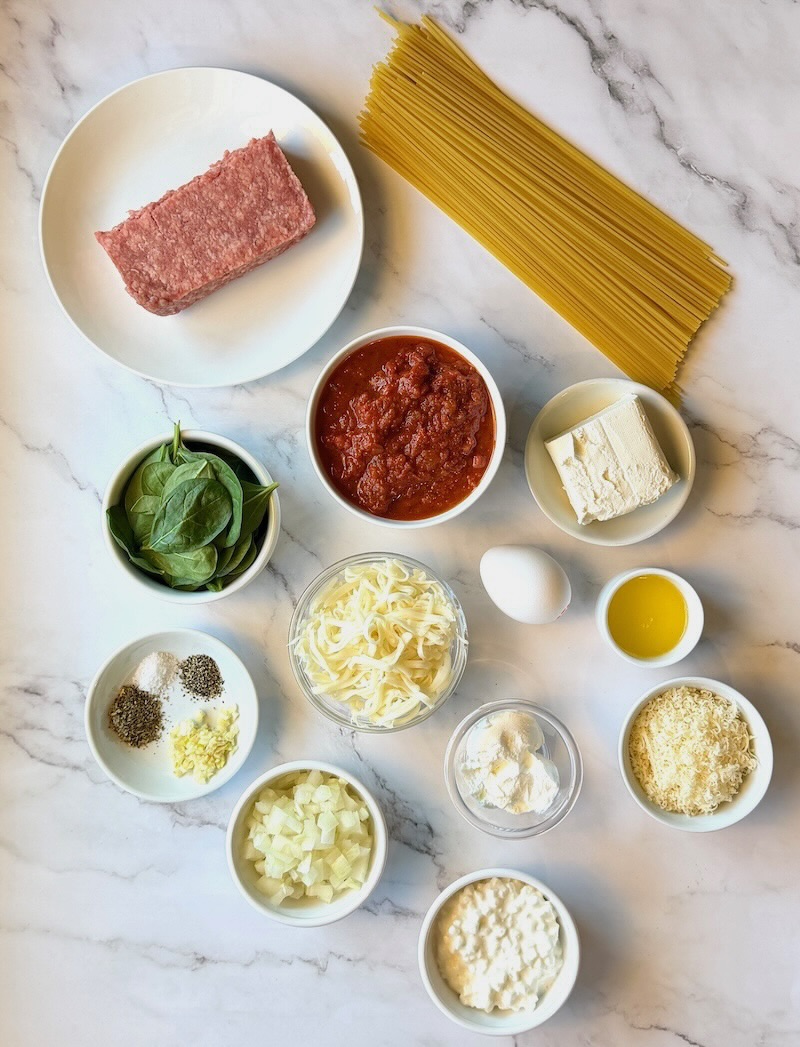 Ingredients displayed for million dollar baked spaghetti. ground beef, onions, garlic, seasonings, noodles, pasta sauce, cream cheese, sour cream, cottage cheese, egg, butter, parmesan cheese, mozzarella cheese, and spinach.