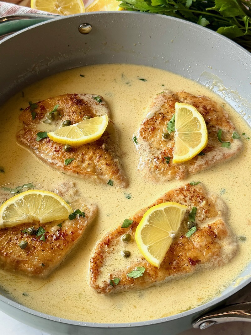 Chicken piccata in a skillet with cream sauce and topped with fresh lemon slices and parsley. 