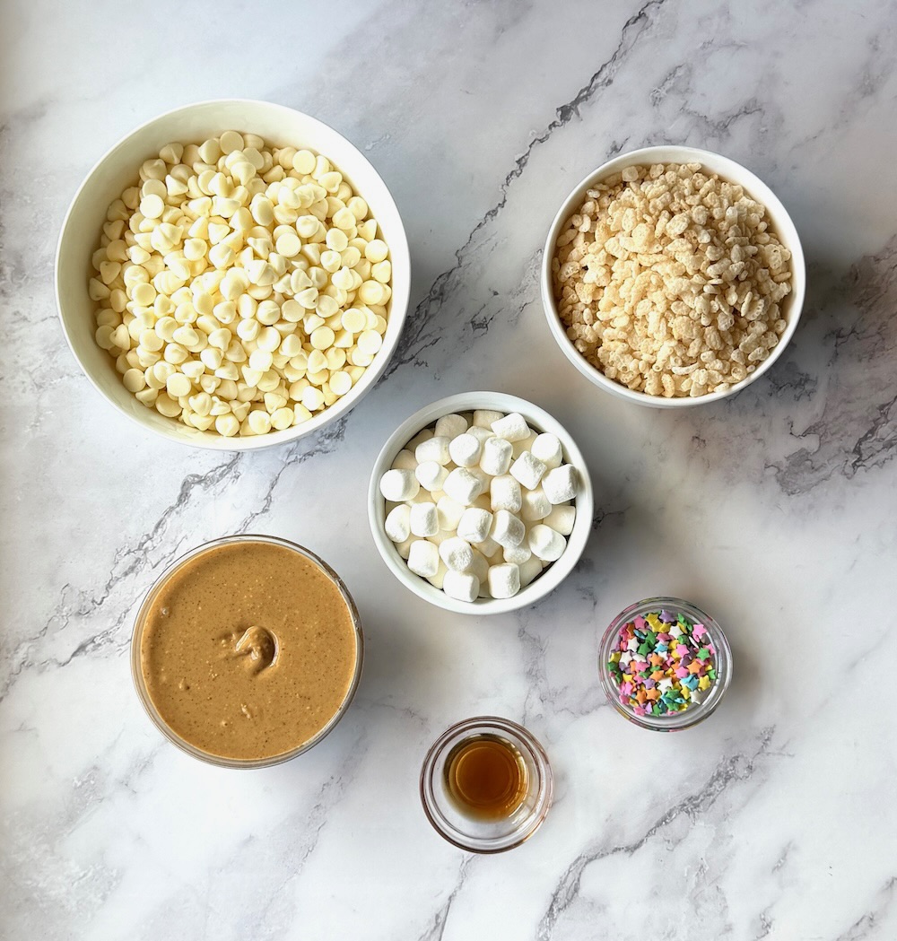 Ingredients for avalanche cookies. Rice krispies, white chocolate chips, creamy peanut butter, vanilla extract, mini marshmallows, and colorful sprinkles.