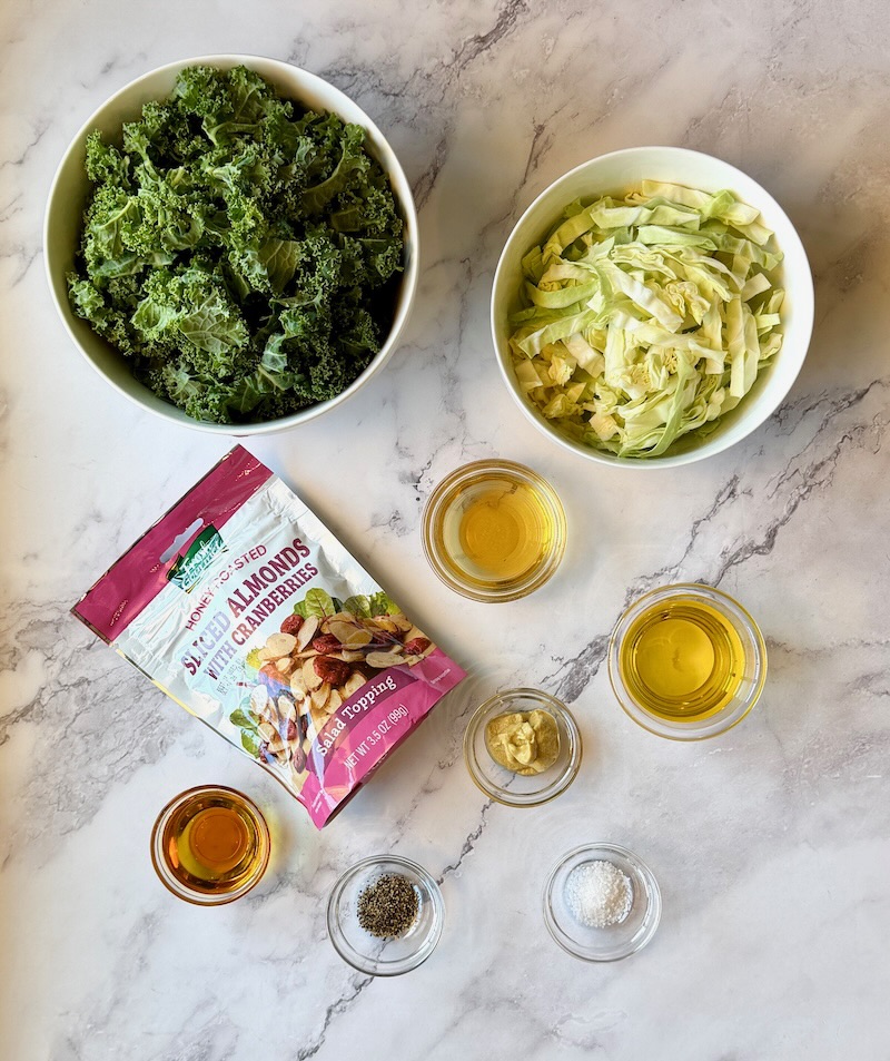 Ingredients displayed for easy kale crunch salad. Kale, green cabbage, almonds and cranberries, olive oil, apple cider vinegar, maple syrup, Dijon mustard, salt and pepper. 