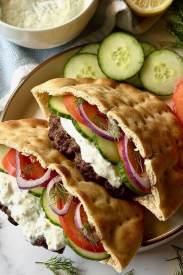 Greek pita burgers assembled on a plate with cucumbers and tomatoes.