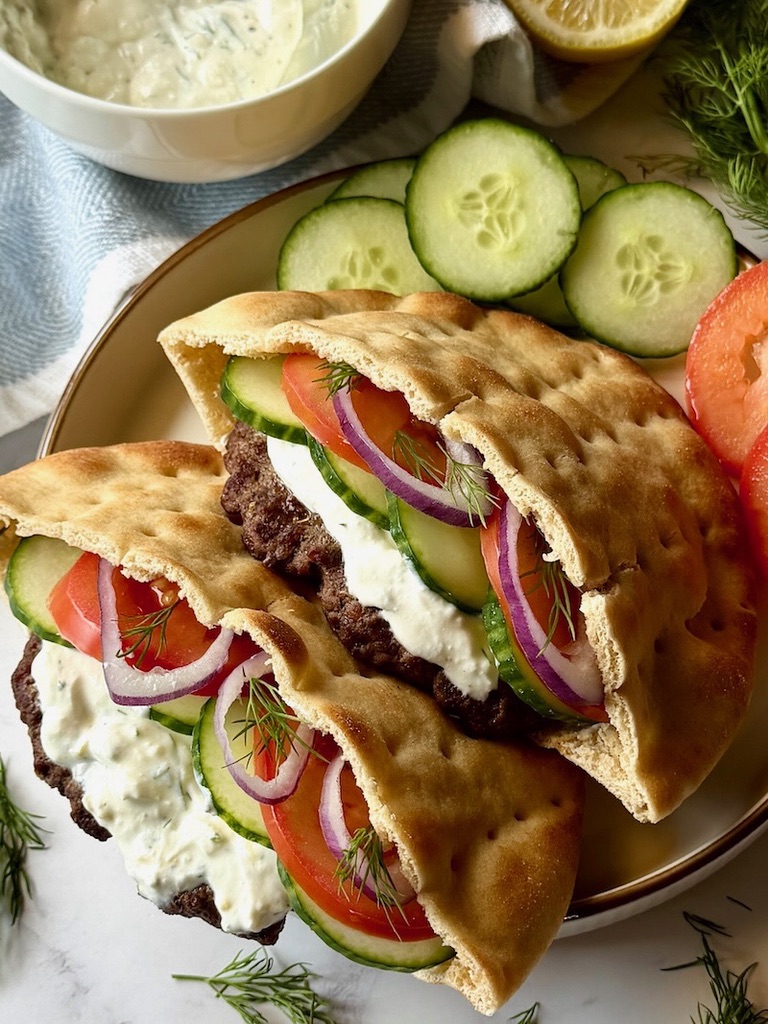Greek pita burgers assembled on a plate with cucumbers and tomatoes.